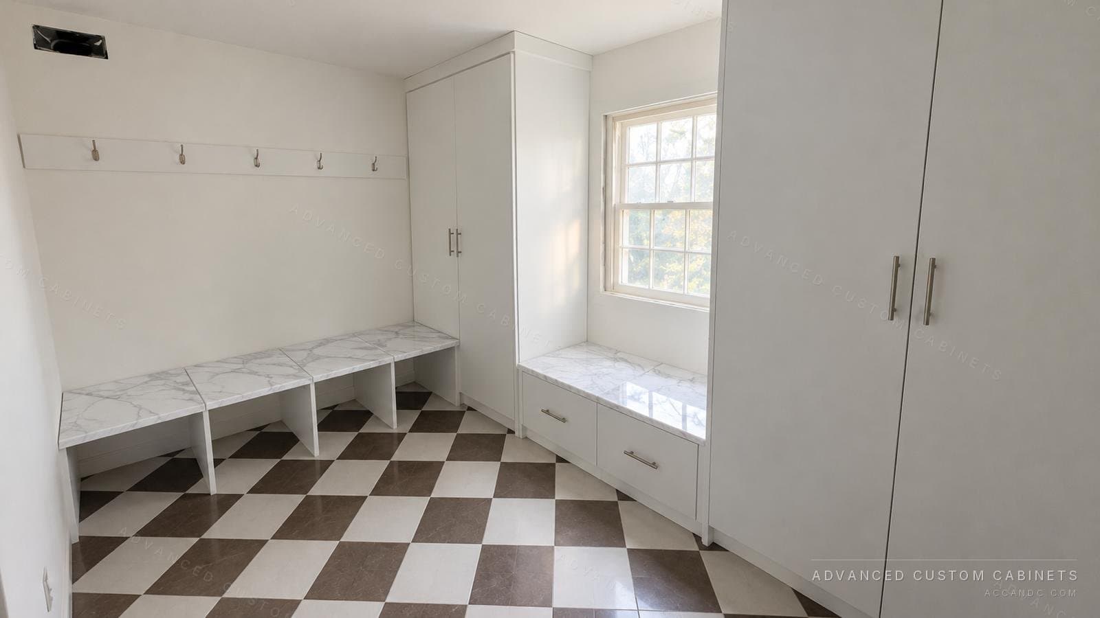 White mudroom with a marble-top bench under a coat hook rail, tall flat-panel cabinets and a window seat with drawers over a checkerboard floor