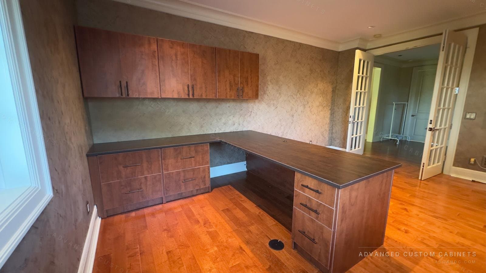 L-shaped home office desk in warm cherry-tone wood with a dark charcoal top, two file-drawer bases, a three-drawer pedestal and matching wall cabinets above, on a hardwood floor beside French doors