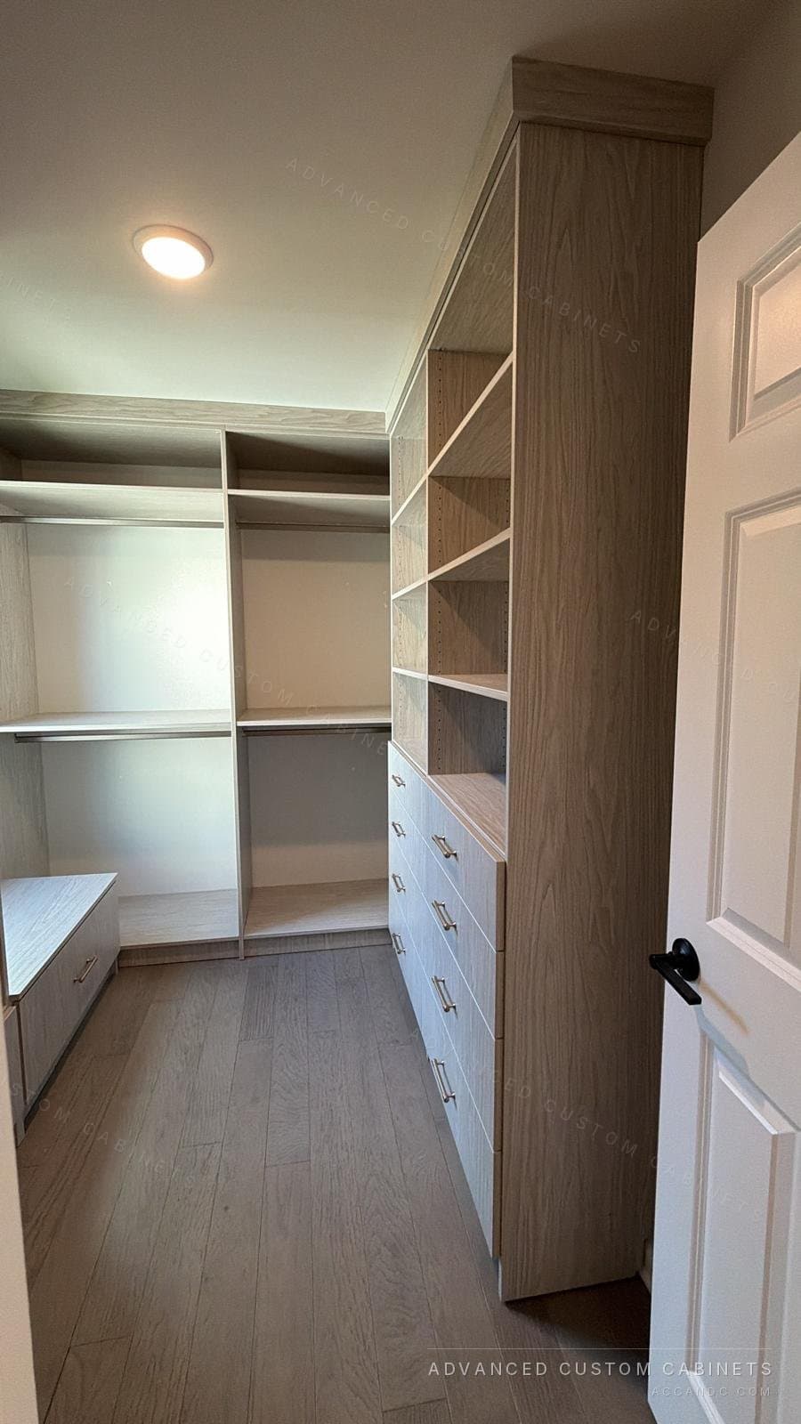 Grey-oak walk-in closet with a tall drawer tower, open shelving and hanging sections beside the entry door