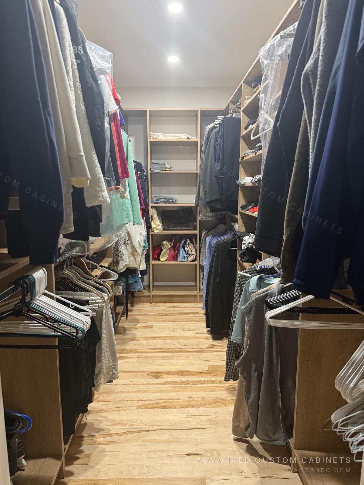 Wood-finish walk-in closet in daily use, with hanging sections on both sides and a shelving tower for folded clothes and handbags at the end