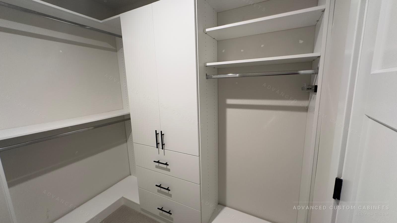 Wide view of a white walk-in closet with a door cabinet over drawers in the center and hanging rods with shelves on both sides