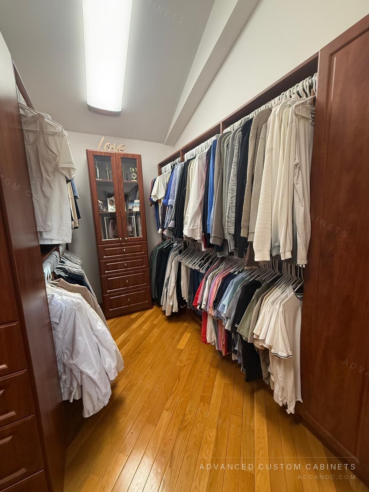 Cherry-finish walk-in closet under a sloped ceiling with double hanging down both sides and a glass-door display cabinet over four drawers at the far end
