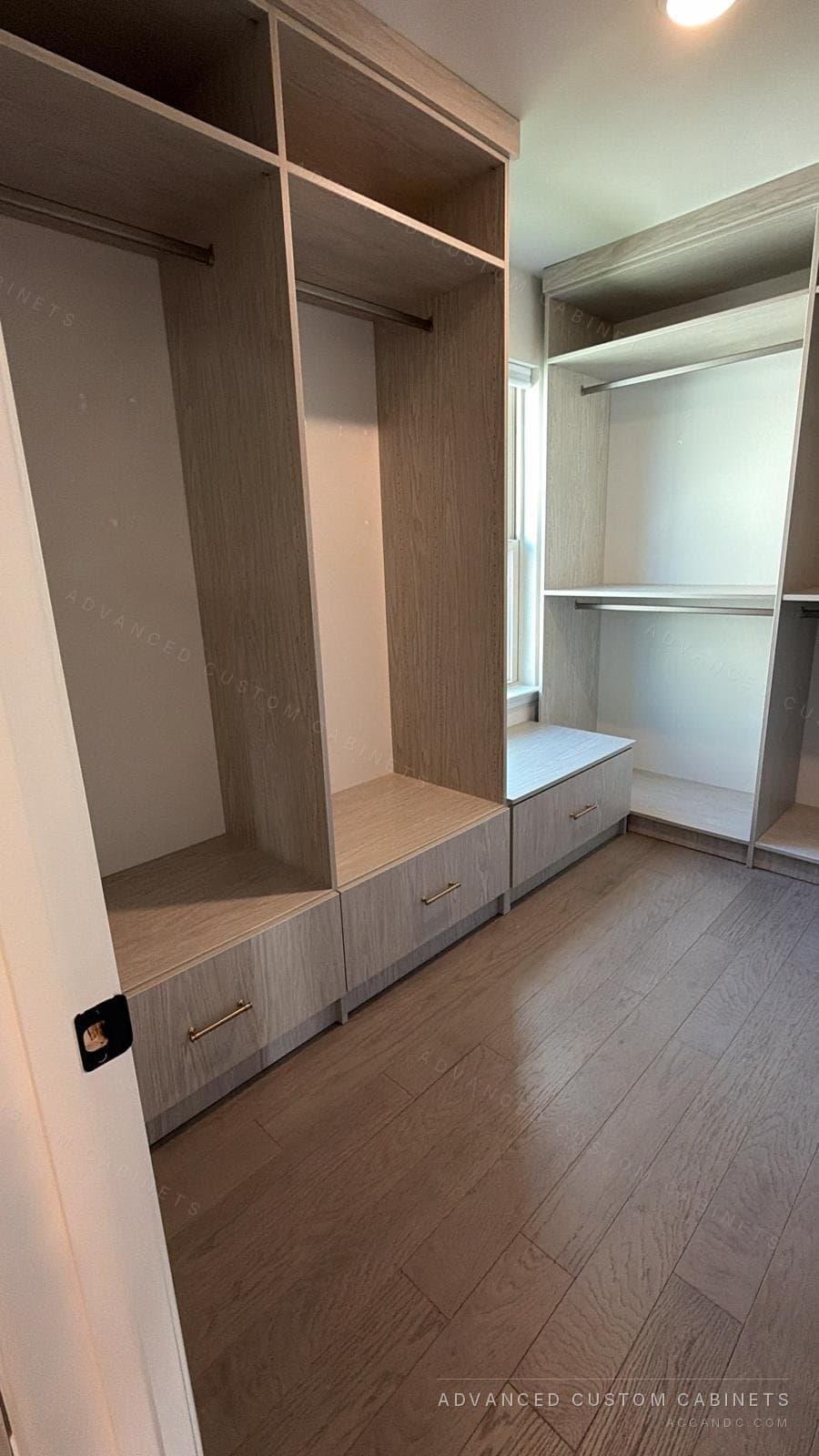 Grey-oak walk-in closet with an L-shaped run of hanging sections, upper shelves and a drawer bench under the window